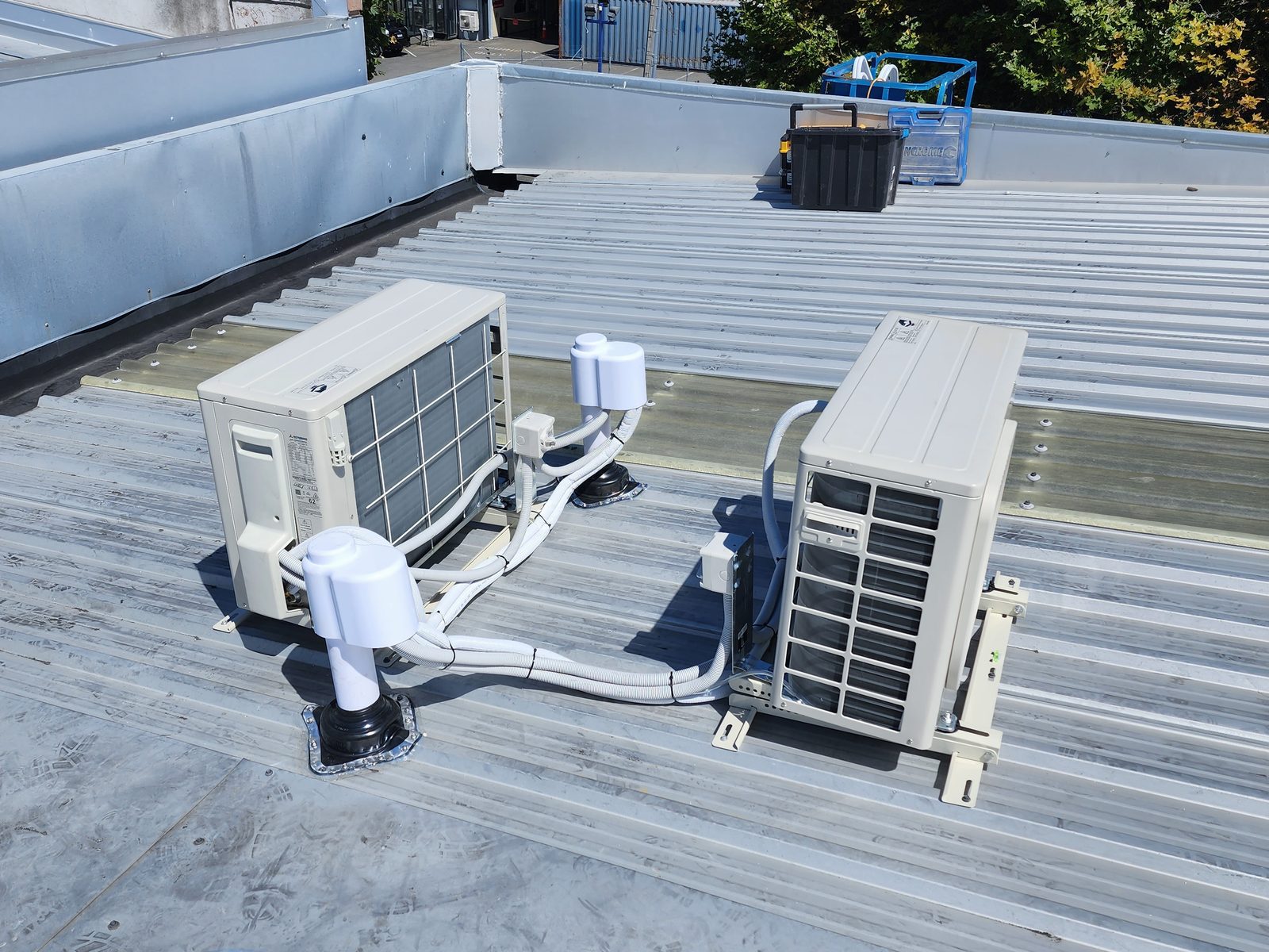 Two outdoor heat pump units on a low-pitch metal roof of a commercial building, with pipework run through roof boots.