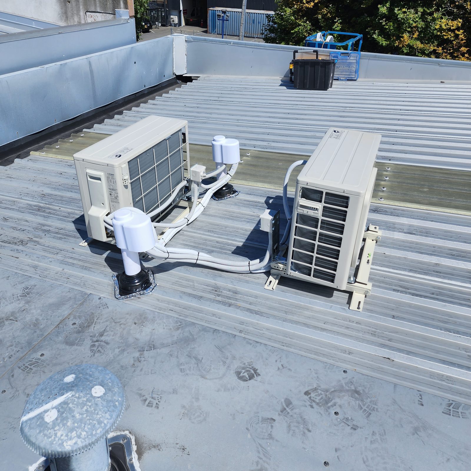Two outdoor heat pump units on a low-pitch metal roof of a commercial building, with pipework run through roof boots.