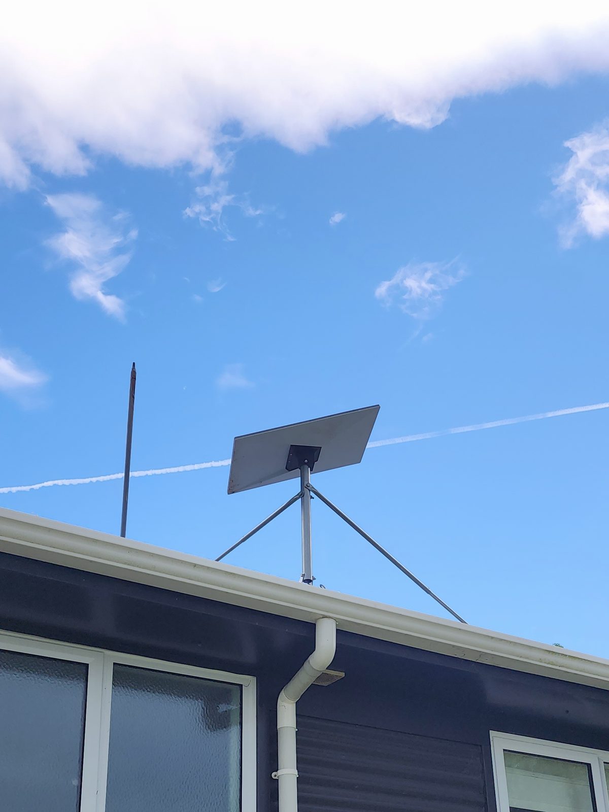 A flat rectangular satellite dish on a tripod mount on the roof of a dark-clad house.
