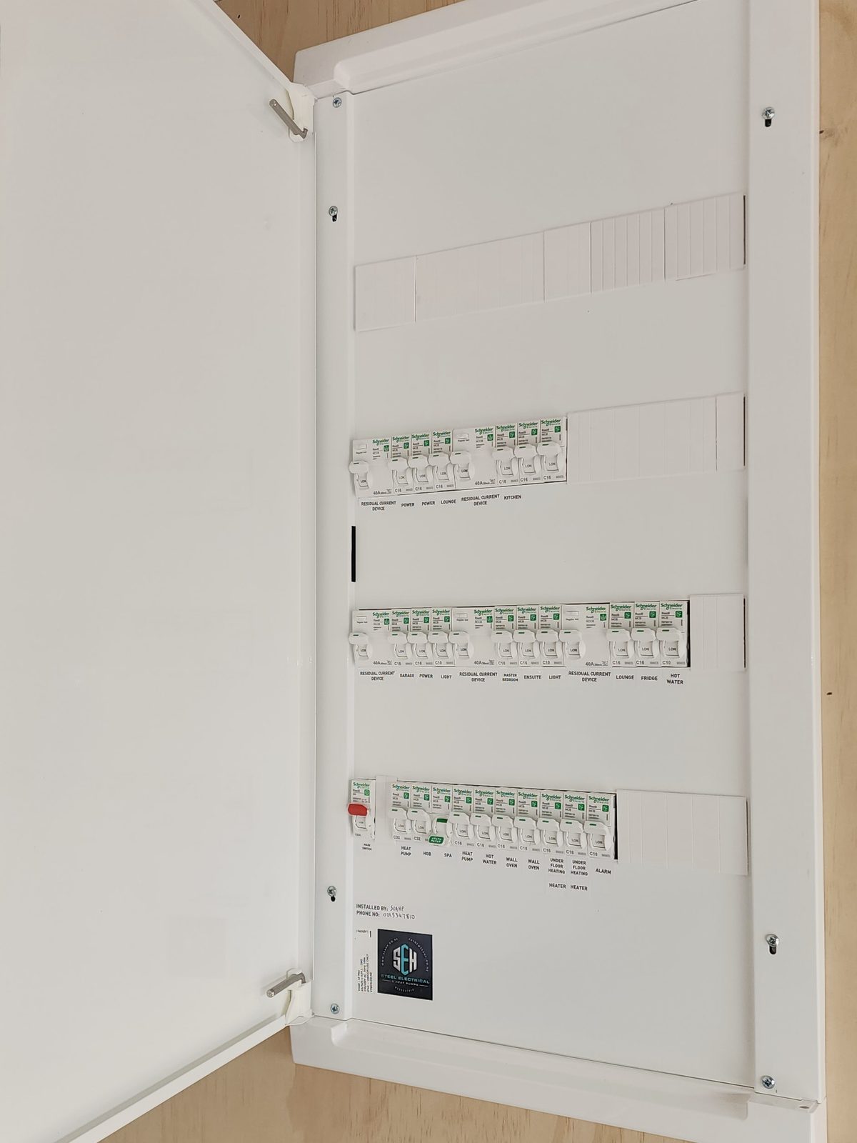 A new white switchboard on a plywood wall with its door open, showing three rows of labelled breakers and a Steel Electrical sticker.