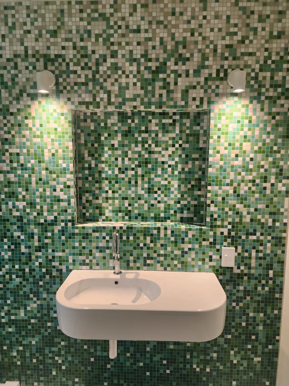 Two white wall spotlights washing a green mosaic-tiled bathroom wall above a wall-hung basin.