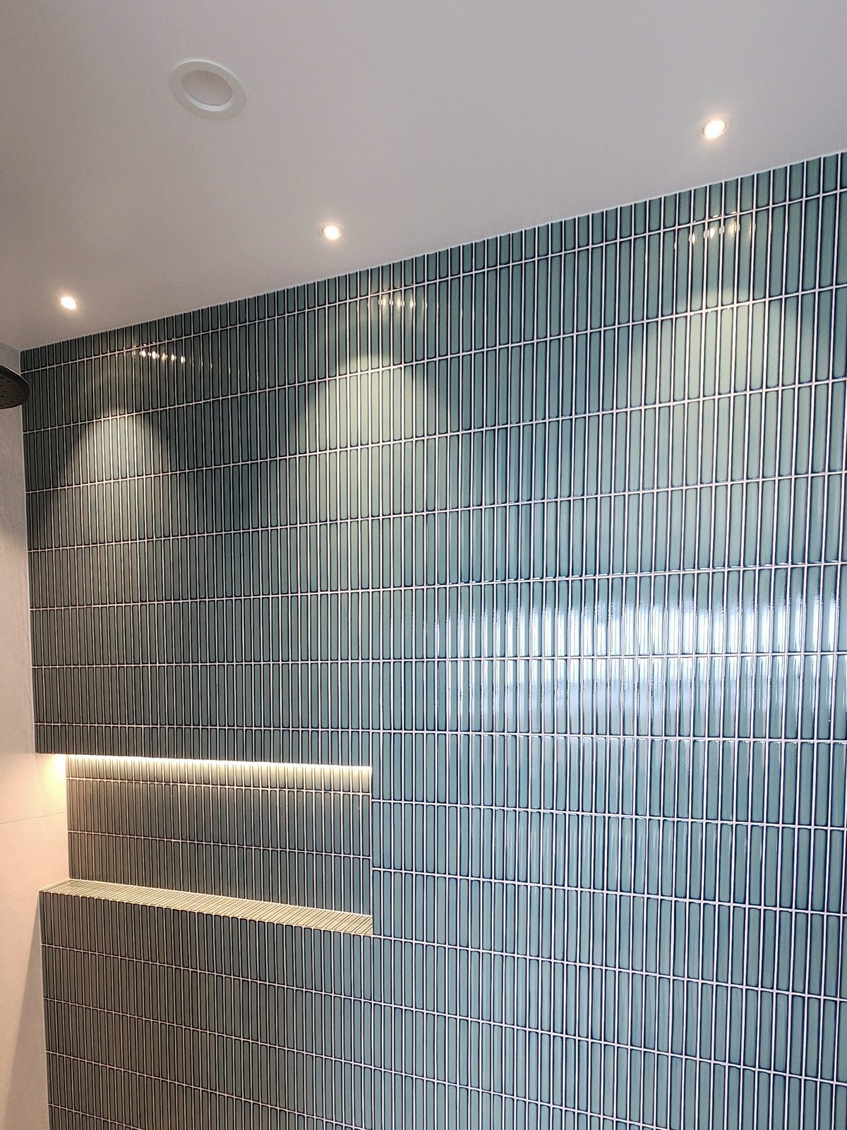 A bathroom wall of teal finger tiles with recessed downlights above and an LED strip lighting a tiled niche.