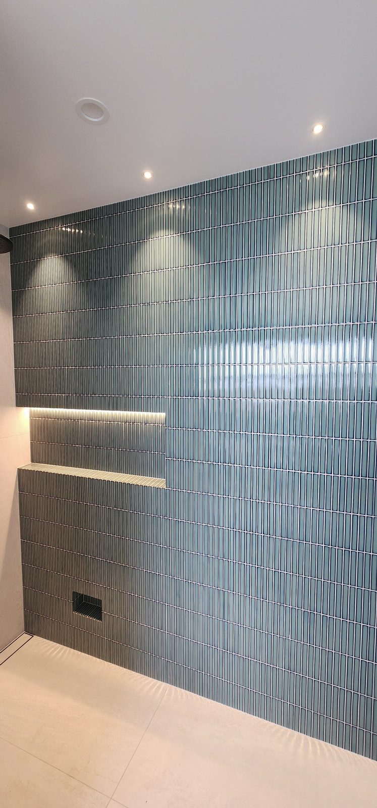 A bathroom wall of teal finger tiles with recessed downlights above and an LED strip lighting a tiled niche.