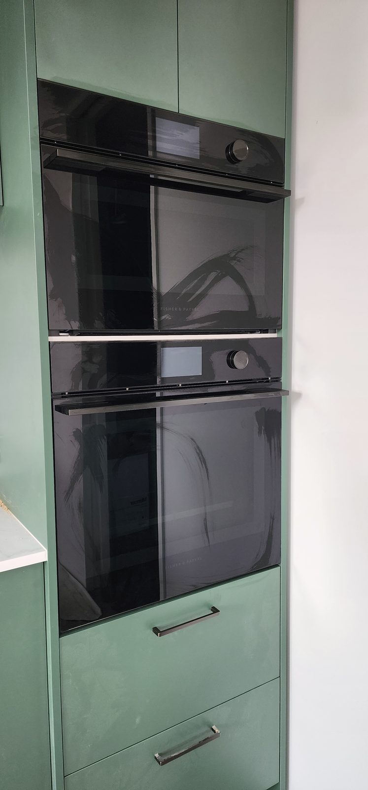 Two black built-in wall ovens stacked in green kitchen cabinetry.