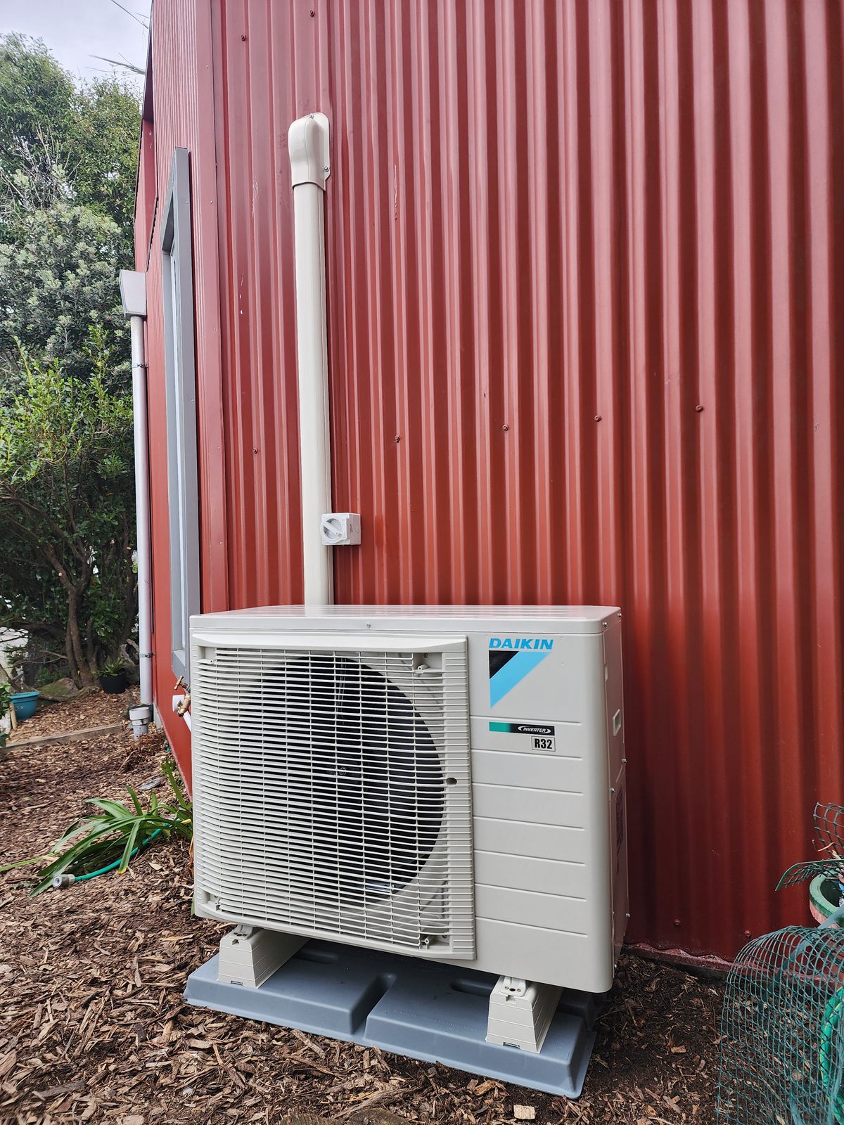 A Daikin outdoor heat pump unit on a plastic base against red corrugated-iron cladding, with capped pipe trunking and an isolator.