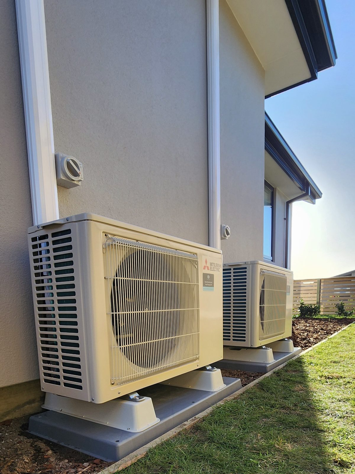 Two Mitsubishi Electric outdoor heat pump units side by side along the plastered wall of a two-storey house, in late sun.