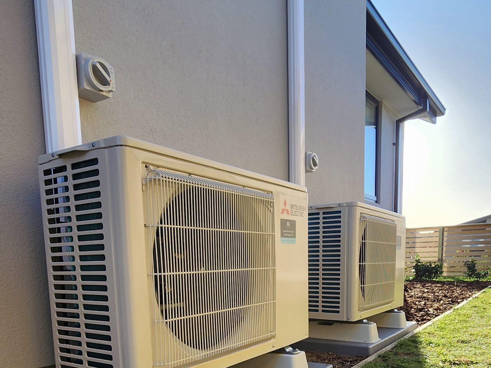Two Mitsubishi Electric outdoor heat pump units side by side along the plastered wall of a two-storey house, in late sun.