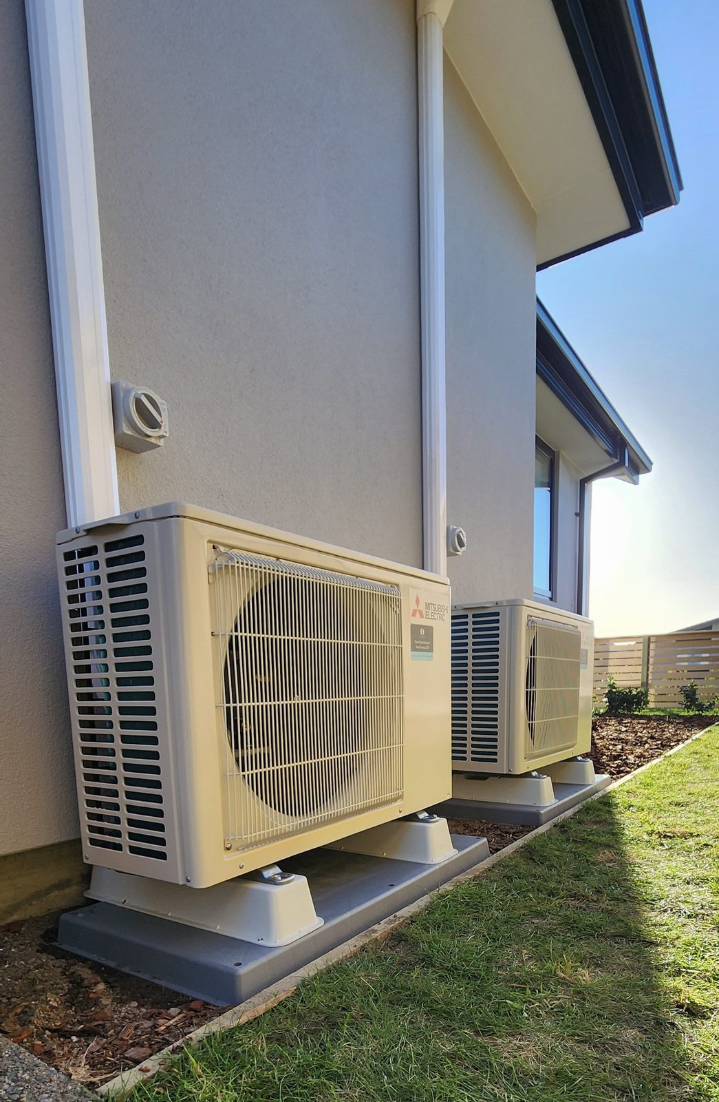 Two Mitsubishi Electric outdoor heat pump units side by side along the plastered wall of a two-storey house, in late sun.
