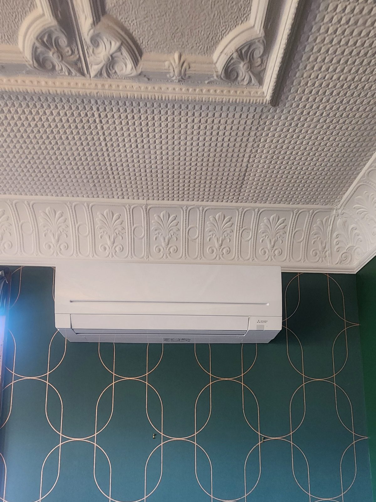 A white wall-mounted heat pump below an ornate pressed-plaster ceiling and frieze, on a dark green patterned wallpaper.