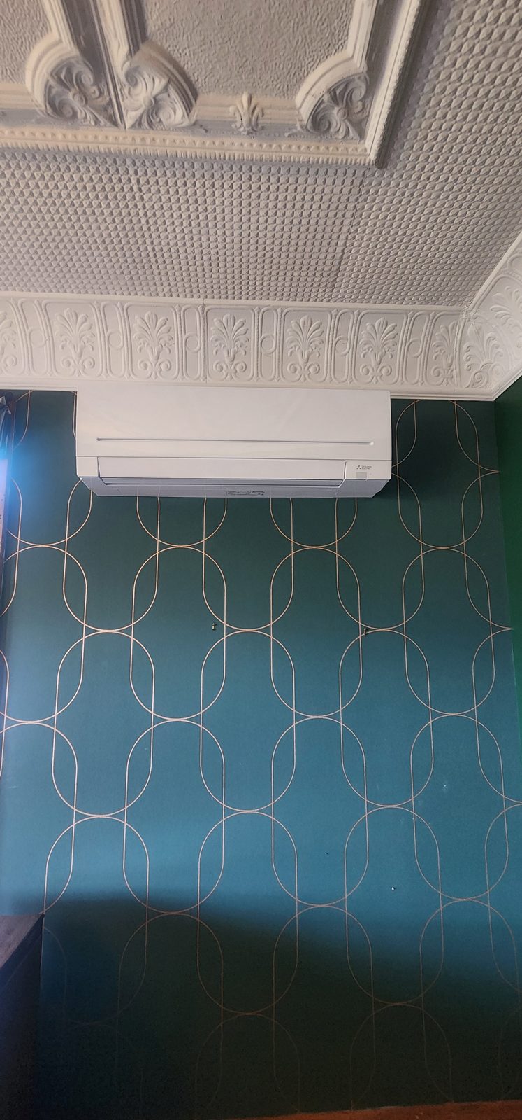 A white wall-mounted heat pump below an ornate pressed-plaster ceiling and frieze, on a dark green patterned wallpaper.