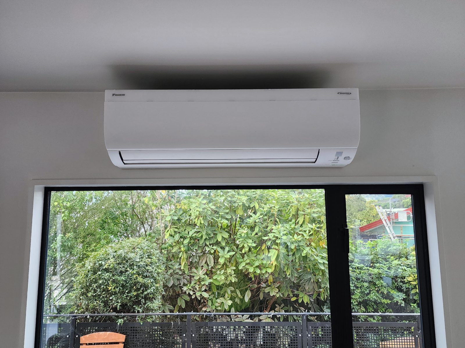A white Daikin wall-mounted heat pump centred above a black-framed window looking onto a planted courtyard.