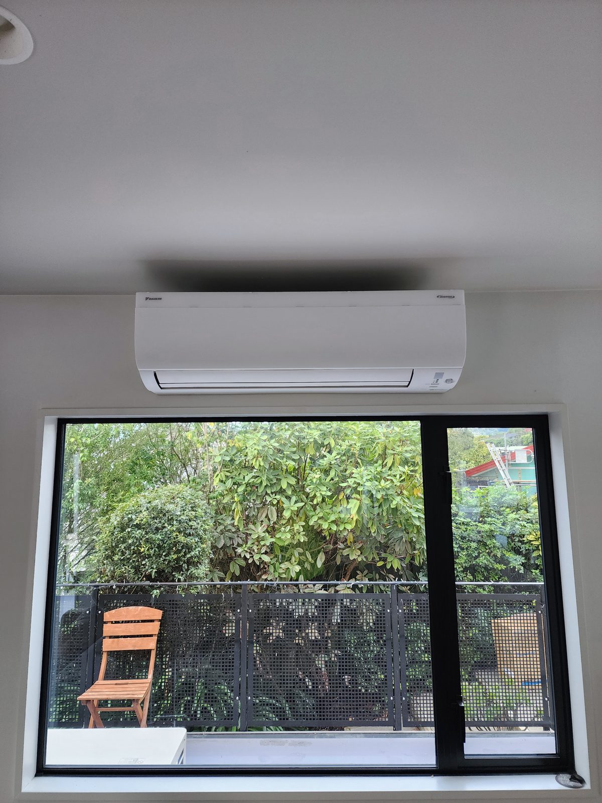 A white Daikin wall-mounted heat pump centred above a black-framed window looking onto a planted courtyard.