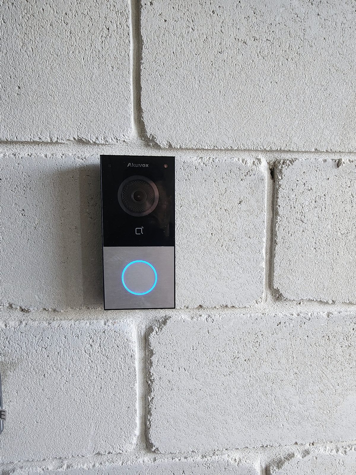 An Akuvox video door station with an illuminated call button on a painted concrete-block wall.