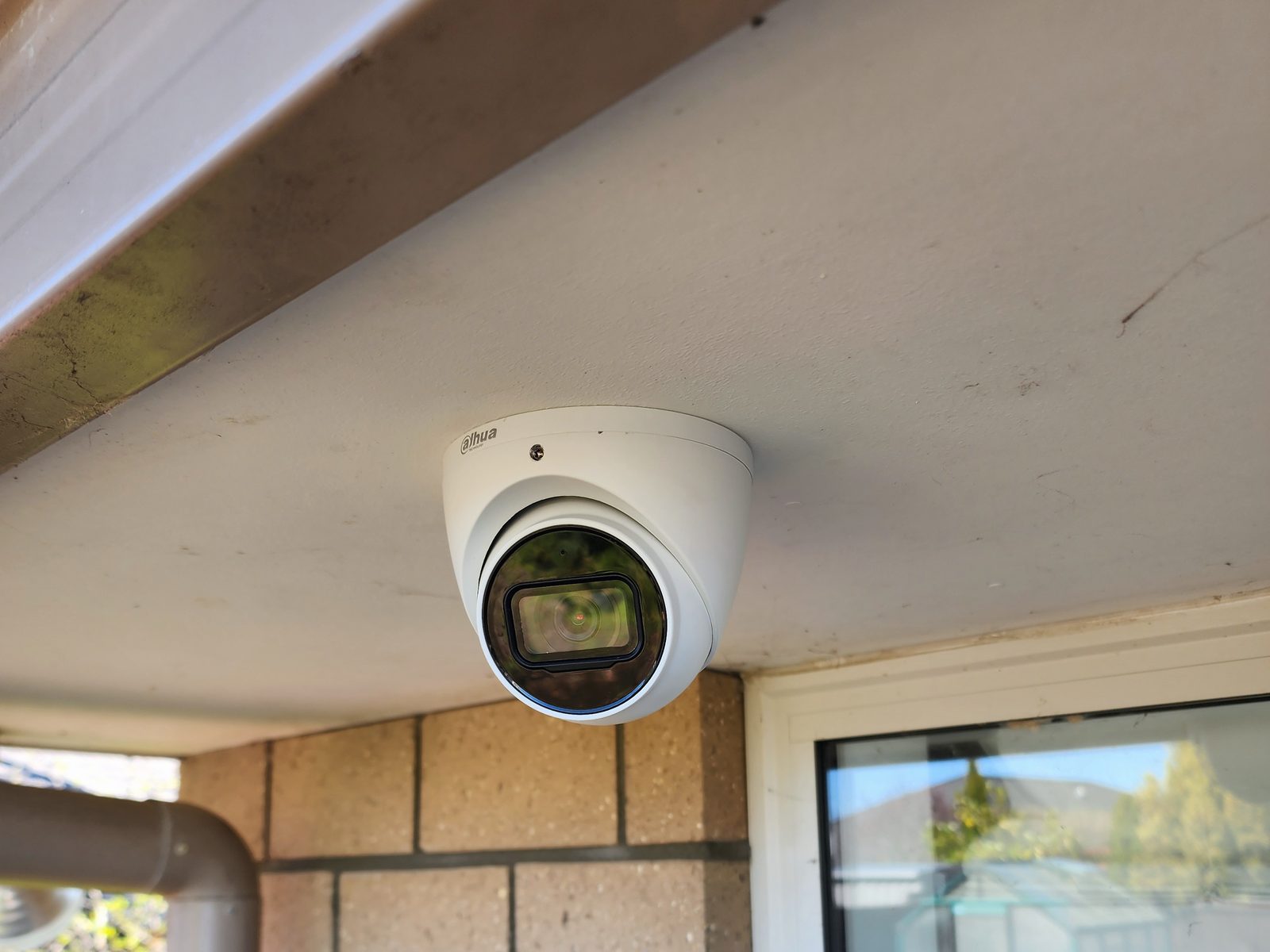A white Dahua turret security camera mounted under the soffit of a brick house.