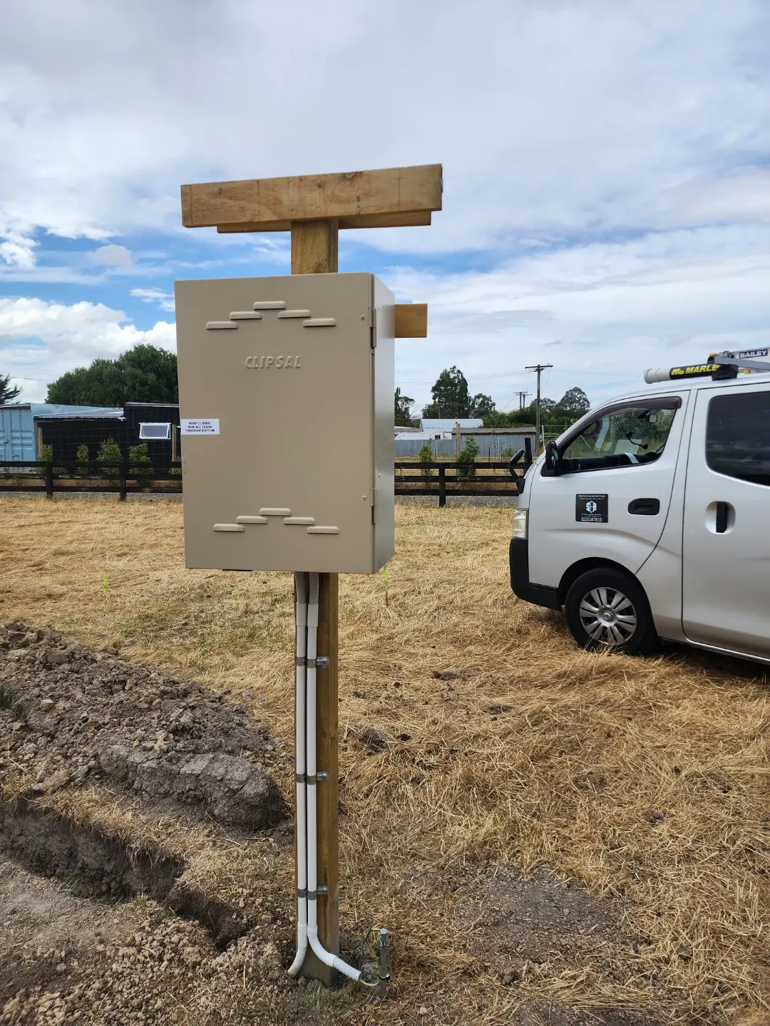 A Clipsal supply pillar on a wooden post in a paddock, with an open trench and conduit at its base and the company van parked behind